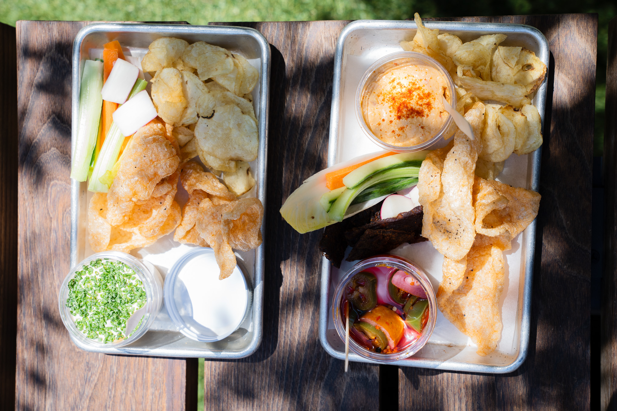 Two trays with chips, pork rinds, veggie sticks, dip, pickled vegetables, and a small salad on a wooden table in sunlight.