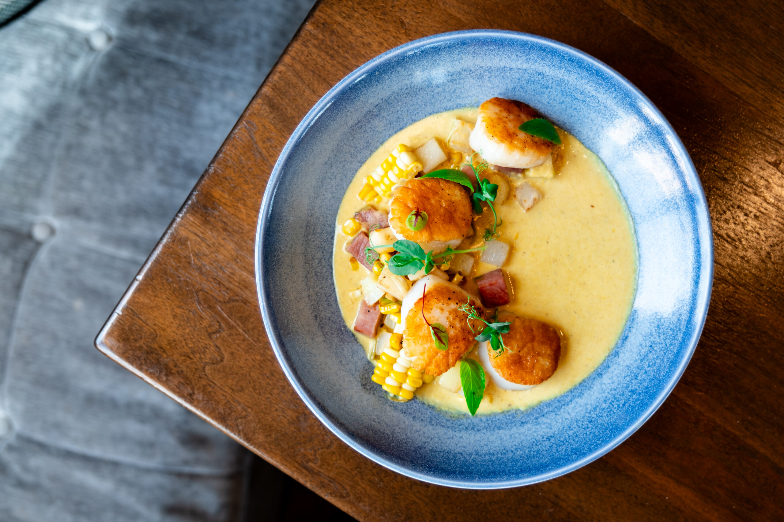 Blue bowl with seared scallops, corn, diced potatoes, and herbs in a creamy yellow sauce, on a wooden table next to a gray cushion.