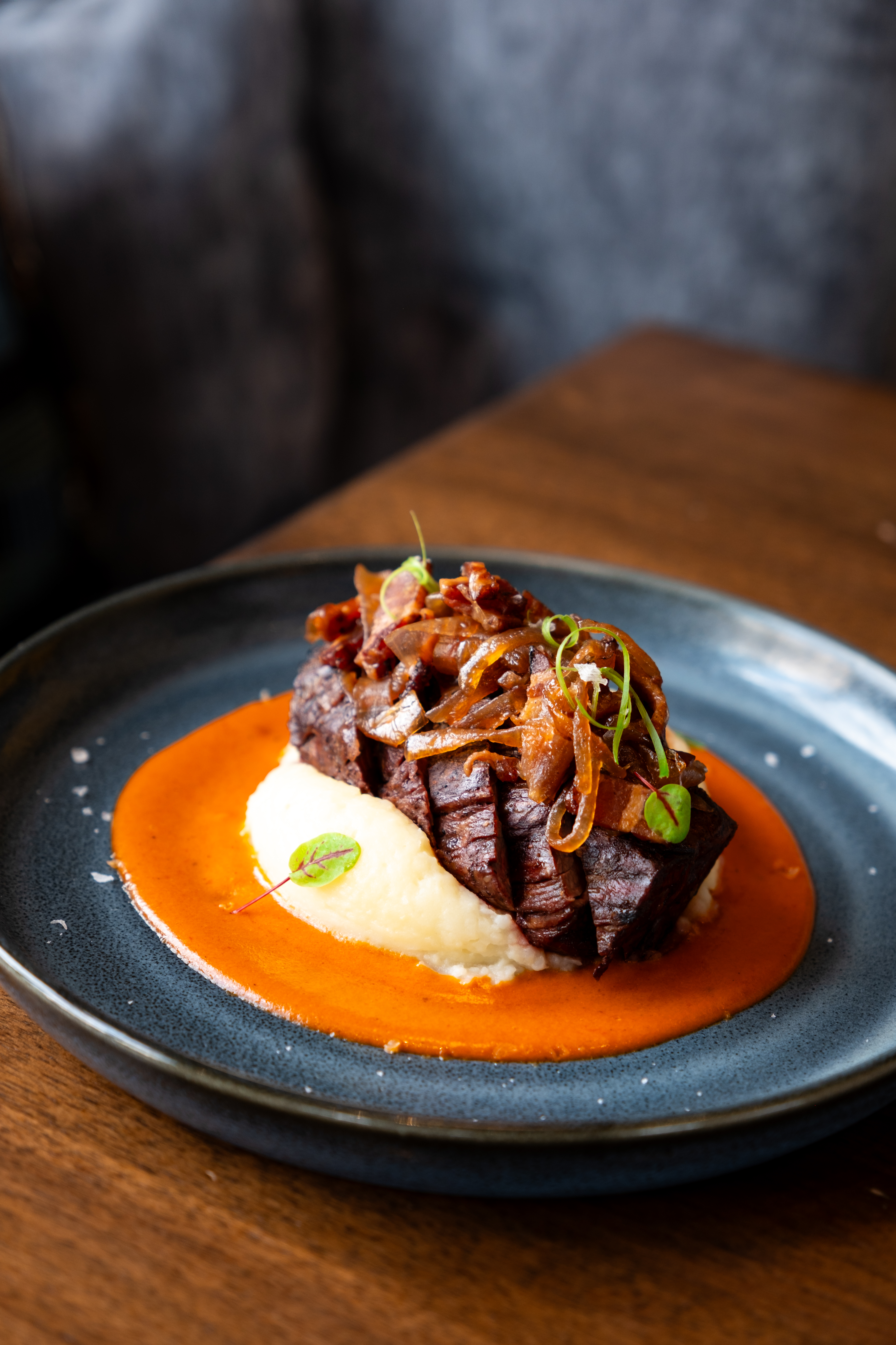 A plate with sliced steak on mashed potatoes, topped with caramelized onions, served with red sauce, on a dark plate on a wooden table.