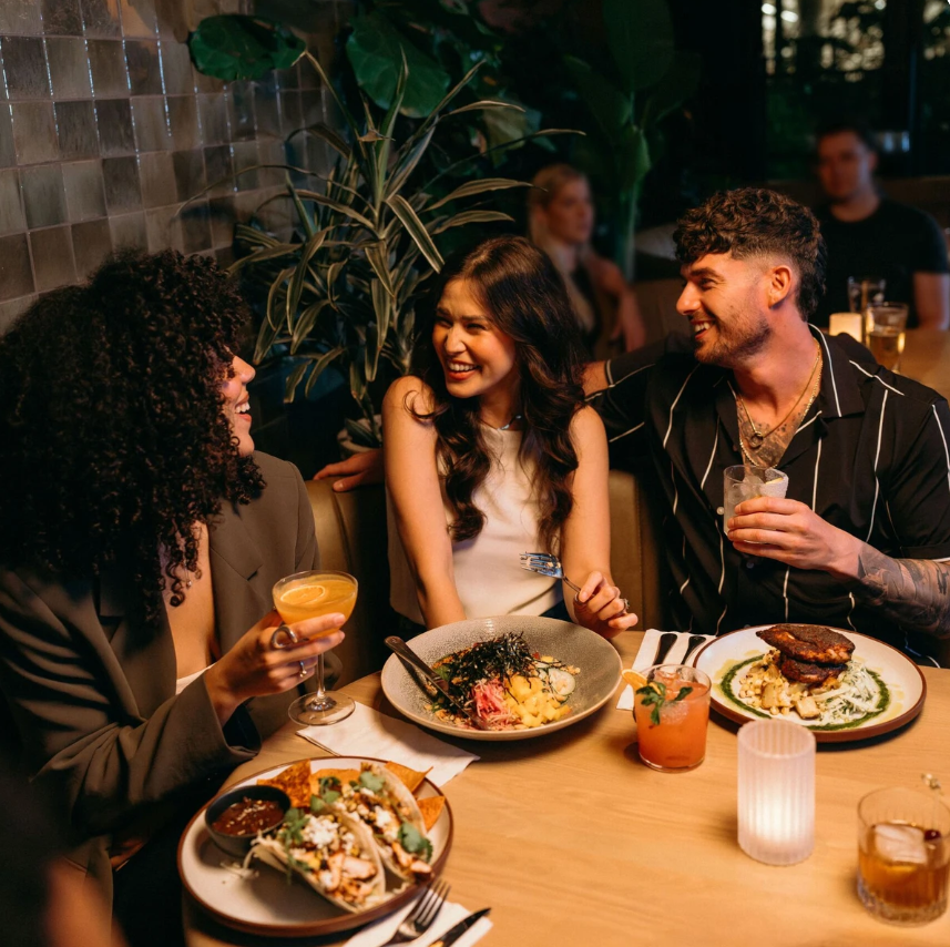 Three friends sit at a restaurant table, smiling and talking, with drinks and plated food in front of them.