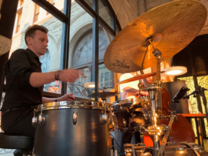 A drummer in a black shirt plays a drum set indoors, with large windows and stone walls in the background.