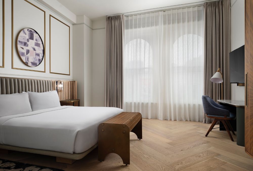 A modern hotel room with a king-size bed, large windows with sheer curtains, a work desk with a chair