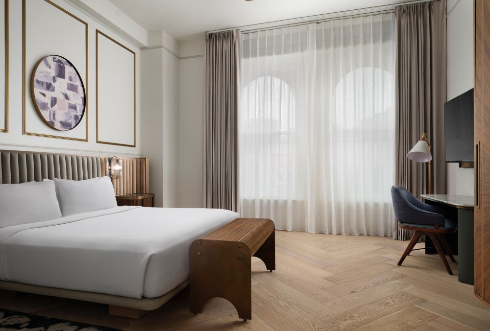 A modern hotel room with a king-sized bed, light-filled windows, and a desk