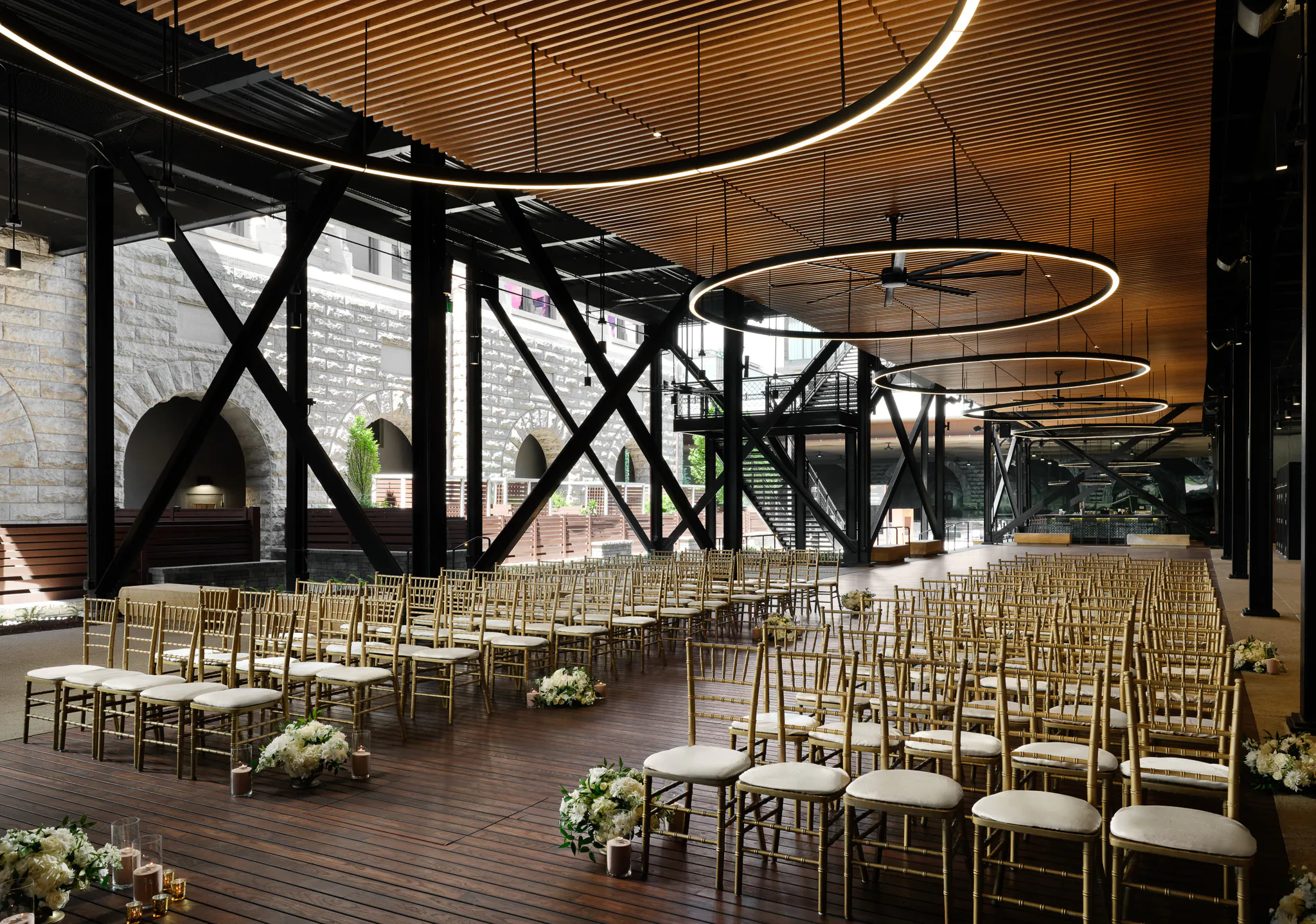 Rows of gold chairs and floral arrangements set up for a wedding ceremony in a modern, open-air venue with circular ceiling lights.