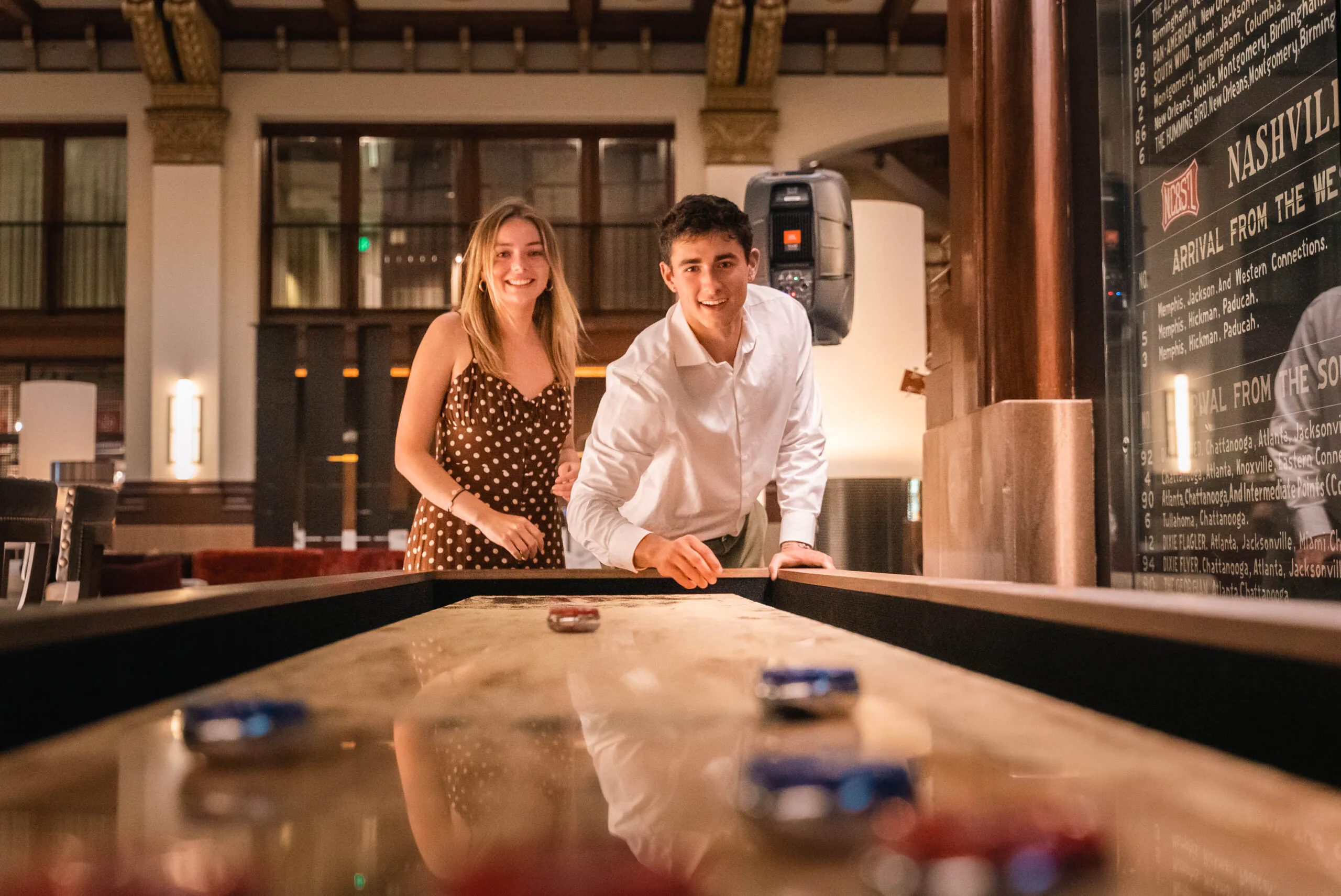 A smiling young woman and man play shuffleboard indoors, with the man mid-shot and game pieces visible on the table.