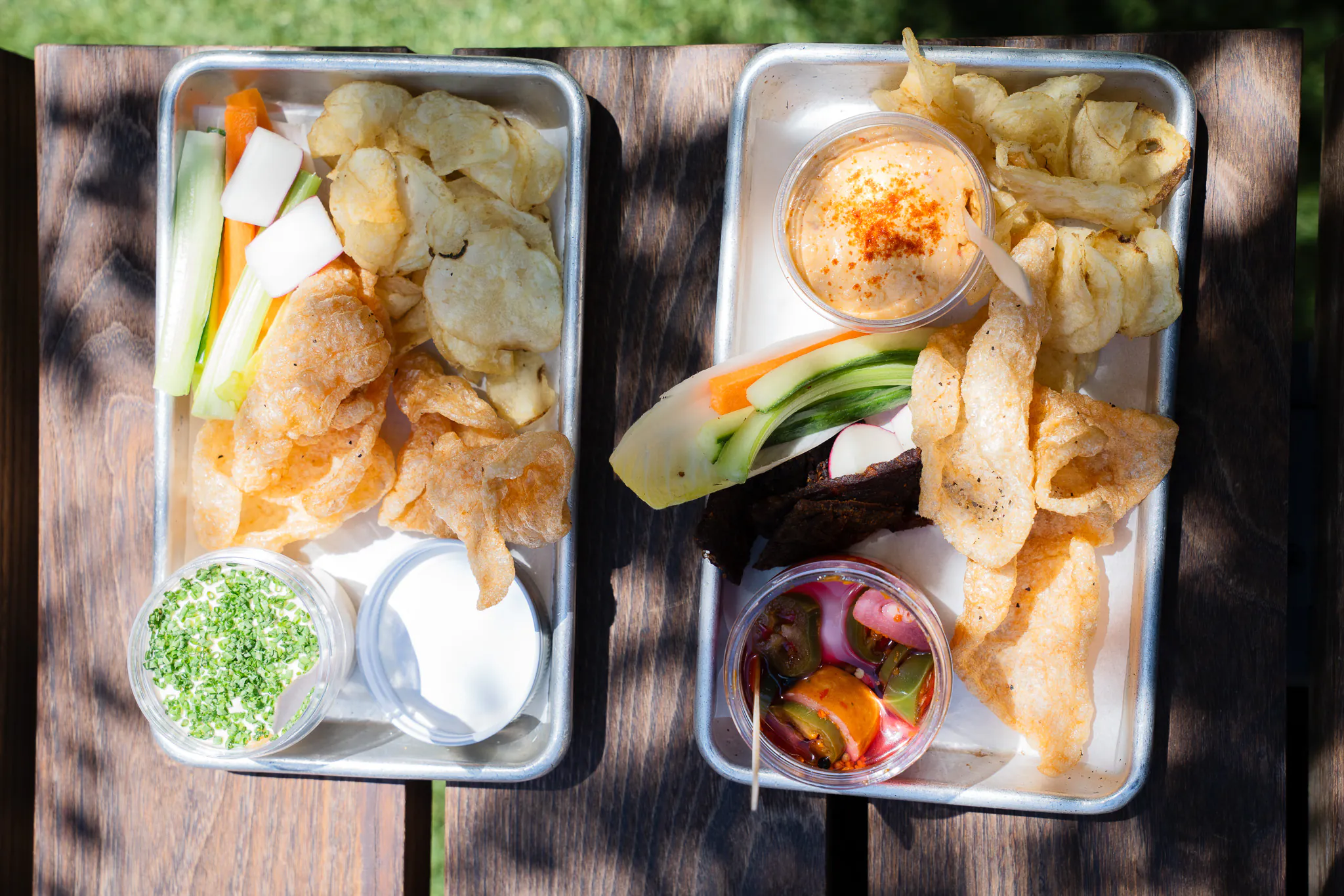Two trays with chips, pork rinds, veggie sticks, dip, pickled vegetables, and a small salad on a wooden table in sunlight.