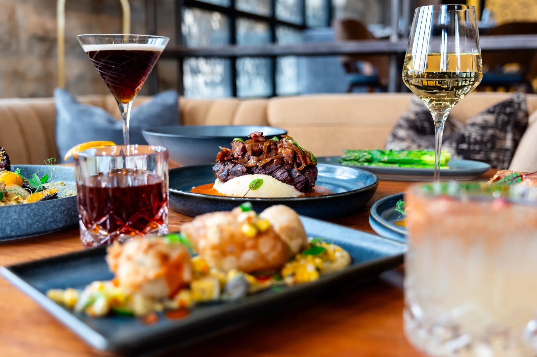 A table set with gourmet dishes, including meat, vegetables, and cocktails, in a stylish restaurant with cozy seating and natural light.