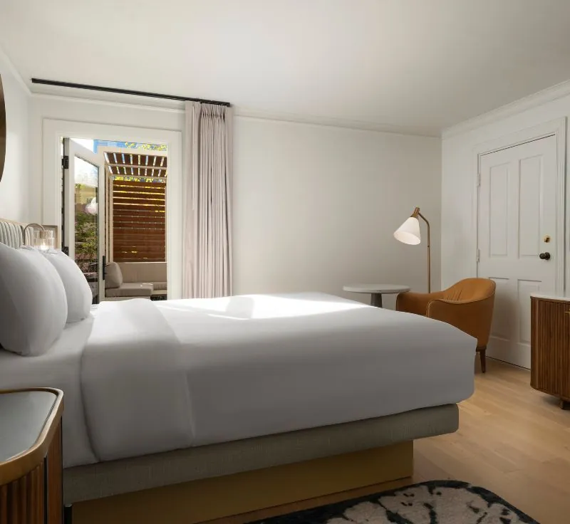 Room with a bed facing a door to a private patio, featuring a small table, chair, and a lamp.