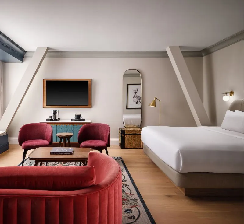 A spacious and modern hotel room with a large bed, red armchairs, and a wall-mounted flat-screen TV.